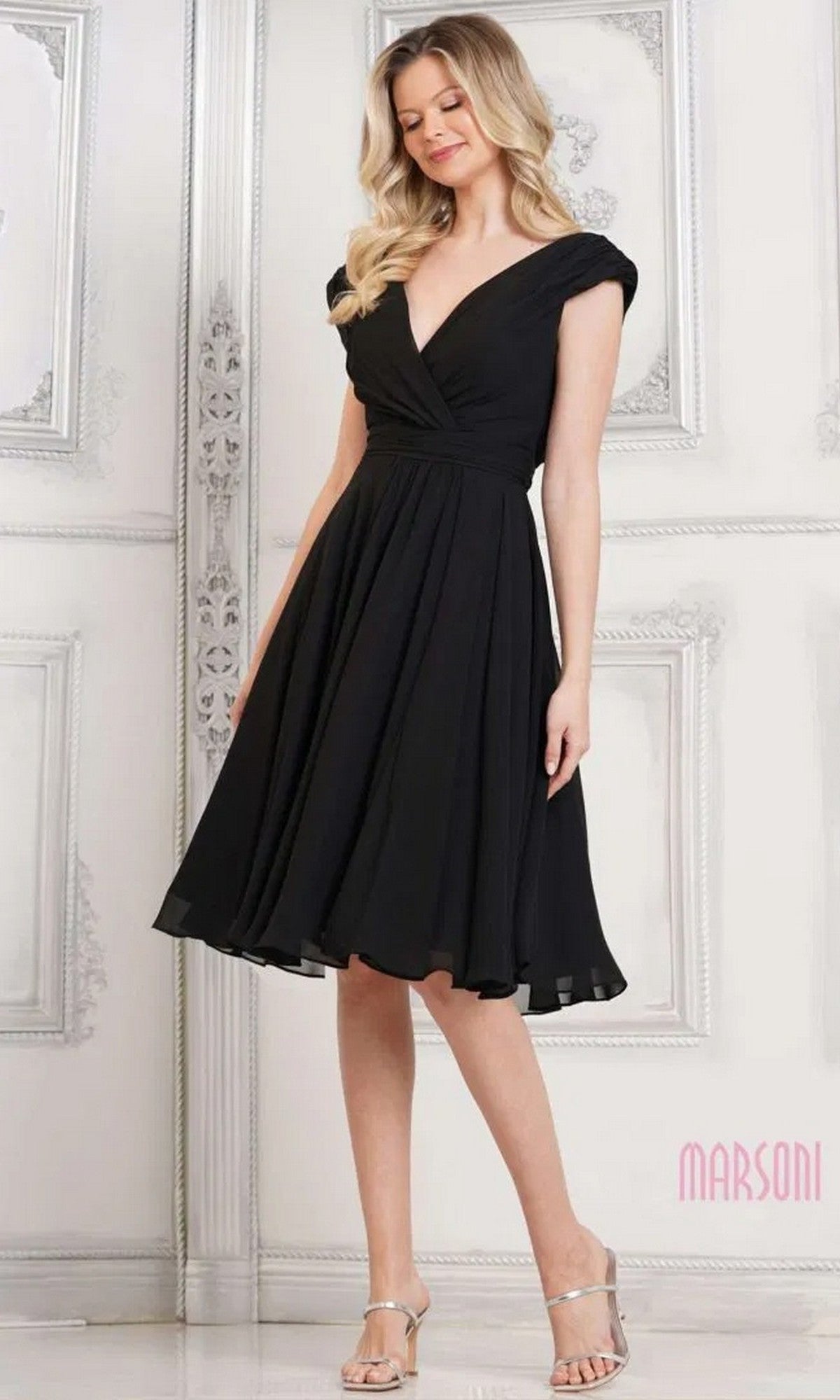 A woman stands against an ornate white wall wearing the Marsoni M251S short semi-formal dress in black, featuring an A-line silhouette, V-neckline, cap sleeves, and paired with silver heels.